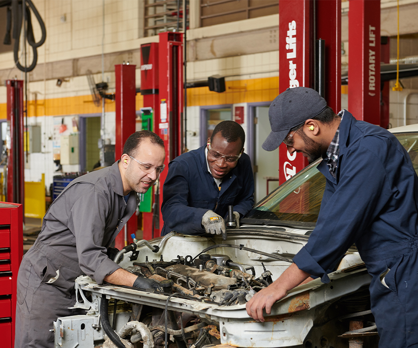 Two students and an Instructor leaning over a cars front end looking at its engine.