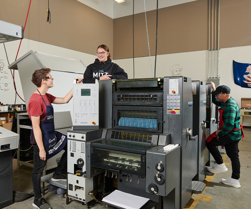 Three students standing around a commercial printing machine.
