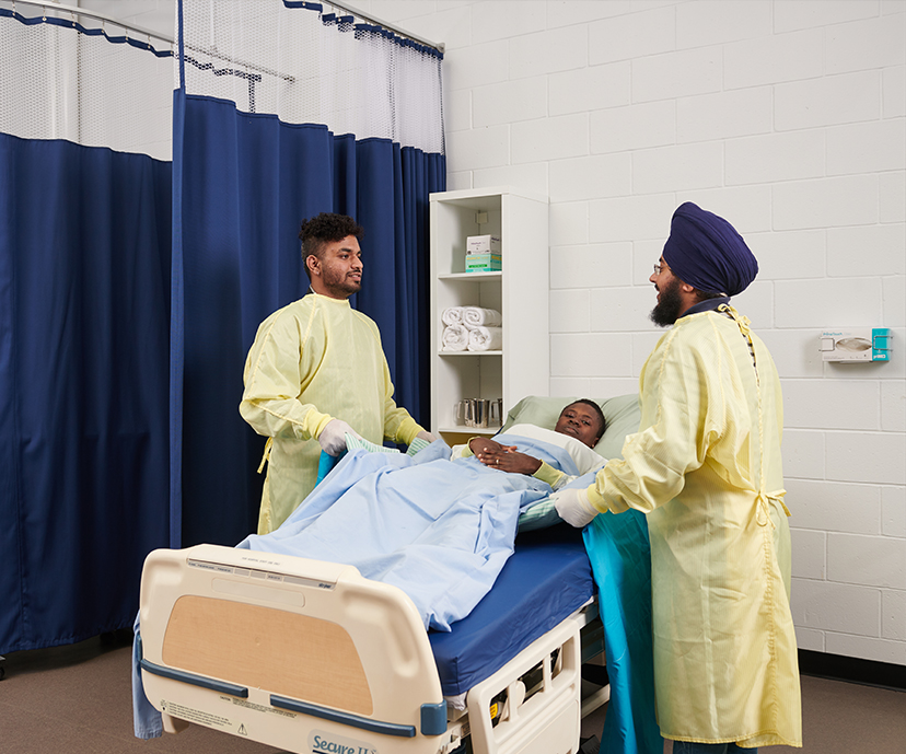 Two Health care students standing over a third student is laying in hospital bed in a mock-up of a hospital room.