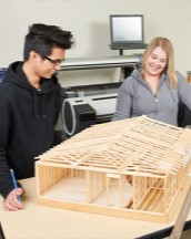 Two students standing over a miniature building make out of wooden sticks.