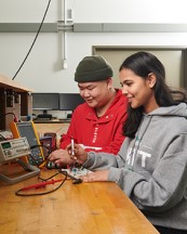 Two students standing at a table using an AMP reader to check the voltage.