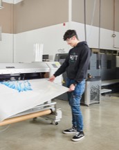 Student standing a wide commercial print machine, waiting for their design to print.