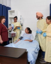Four Health Cares students standing in a mock-up of a hospital room chatting with their teacher.