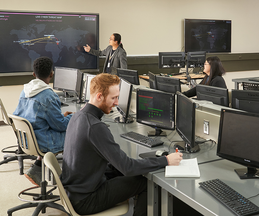 Three students sitting at their desks, in front of them are large computer screens. They are all facing foward looking at the teachers, who is standing in front of a large monitor that is showing that days lesson.